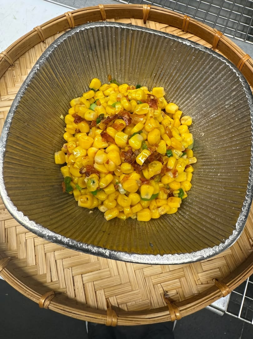 Close-up of a bowl of yellow corn kernels with green onions and brown bits, on a woven mat.