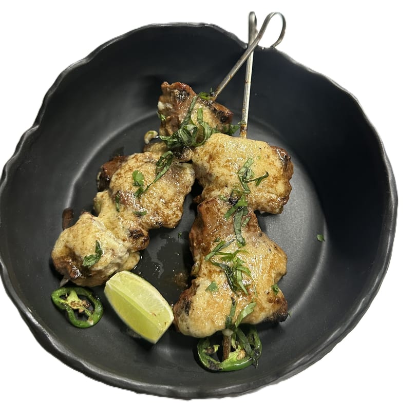 Chicken tikka skewers with creamy sauce, fresh herbs, lime, and jalapeños in a black bowl.
