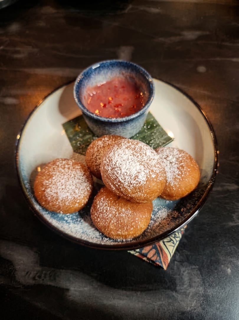 Five powdered sugar-dusted fried dough balls and a small bowl of red dipping sauce on a plate.