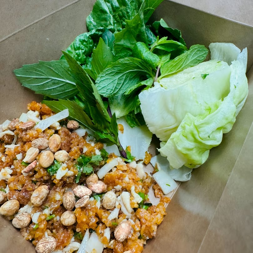 Close-up of a Thai crispy rice salad with peanuts, herbs, and lettuce in a takeout container.