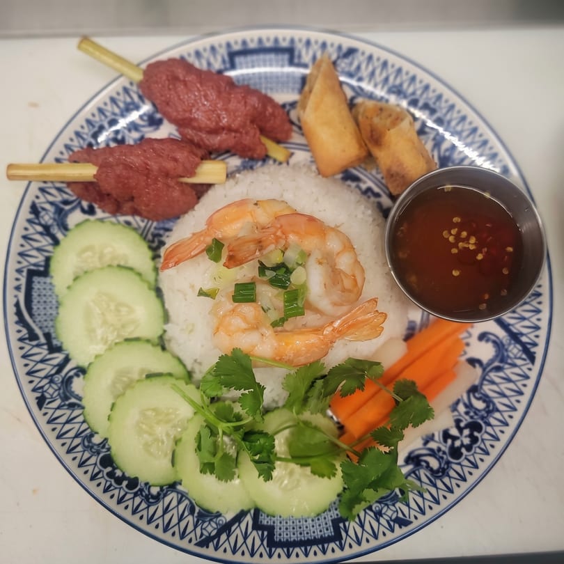 Vietnamese dish with rice, shrimp, cucumber, carrots, spring rolls, and beef skewers on a blue and white plate.